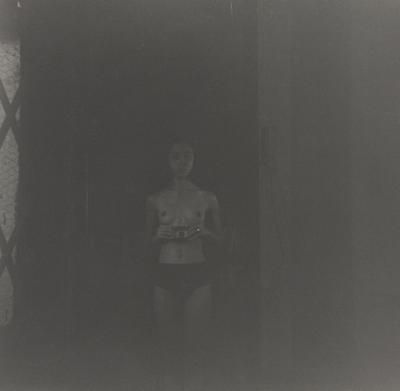 dark black and white image of a woman wearing black briefs holding a camera in front of her belly; small strip panel of chicken wire glass at left with diamond-shaped lattice on top of it; received in silver frame